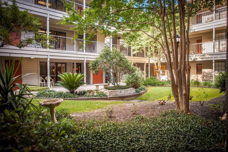 A courtyard garden area within a multi-story residential building featuring green grass, various plants, trees, and a stone birdbath. The building has balconies and doors facing the courtyard, with outdoor chairs visible on the ground floor.