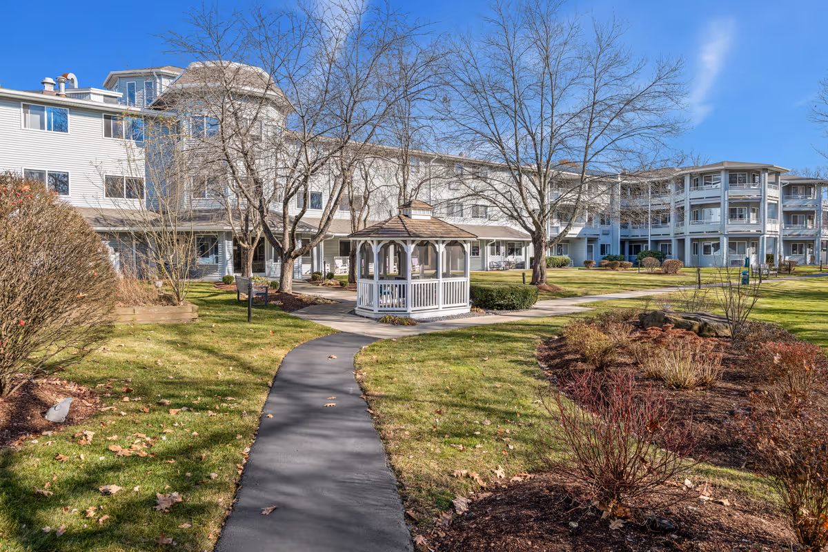 Outdoor courtyard of a senior living community featuring a paved walkway leading to a white gazebo with a multi-story residence in the background.
