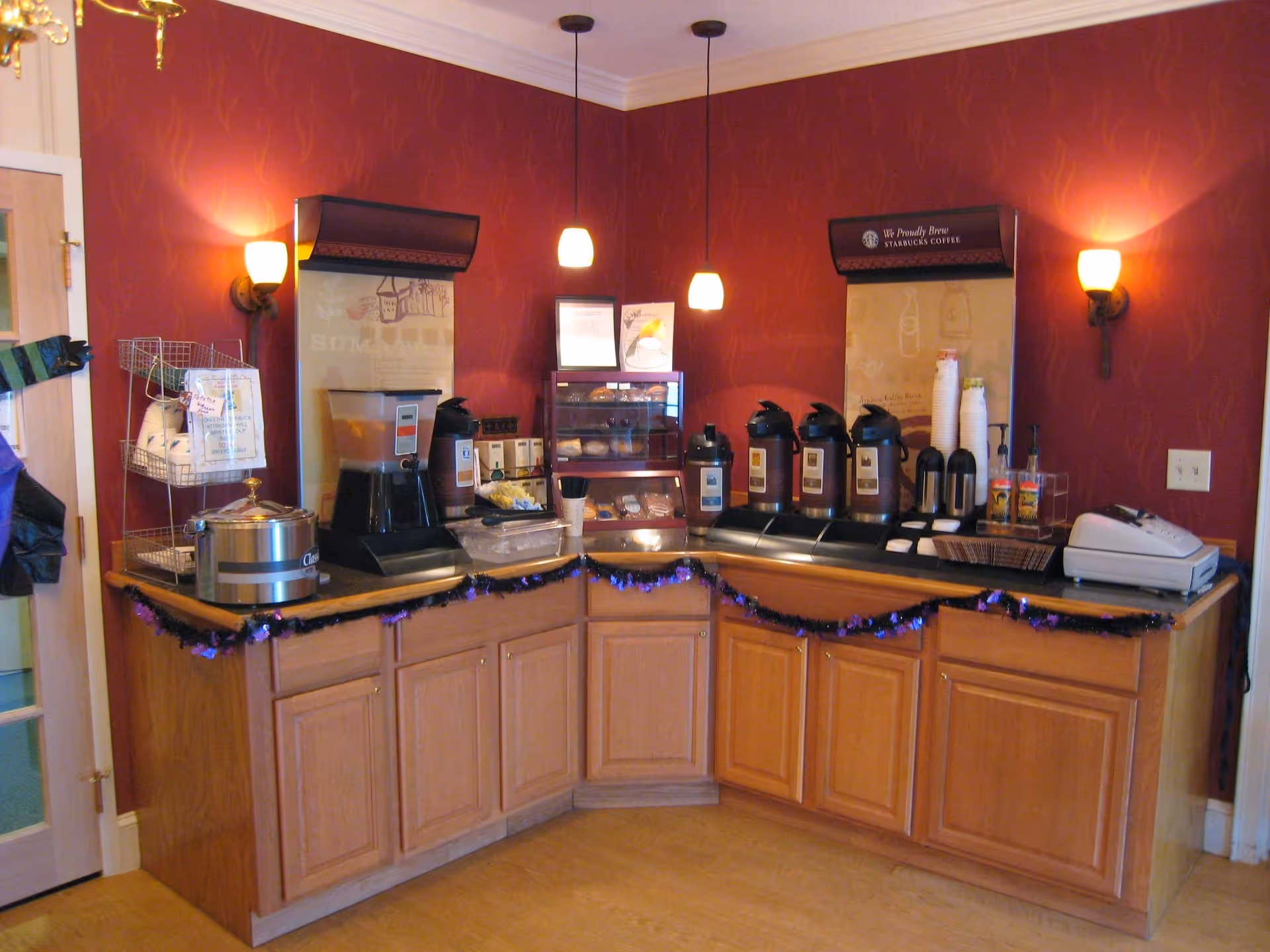 Corner coffee and beverage station with multiple urns, a juice dispenser, cups and cabinets against a red wall.