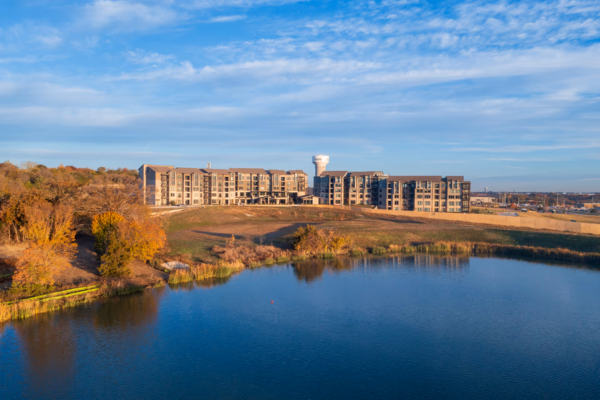 A large multi-story senior living facility building situated on a grassy hill overlooking a calm lake, with trees showing autumn colors and a water tower visible behind the building under a partly cloudy sky.