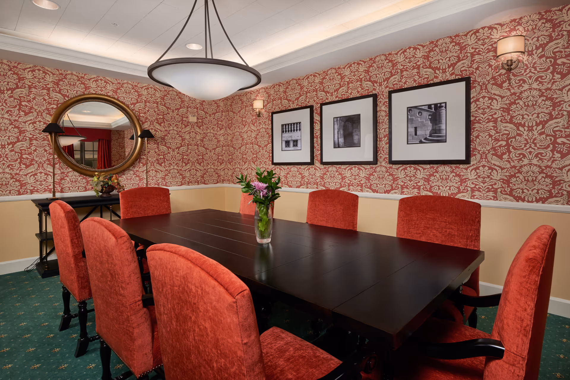 A formal dining room with a dark wooden rectangular table surrounded by eight red upholstered chairs. The walls are decorated with red and beige patterned wallpaper and three black and white framed photographs. A round mirror with a gold frame hangs on one wall above a black console table with a flower arrangement. A large ceiling light fixture illuminates the room.