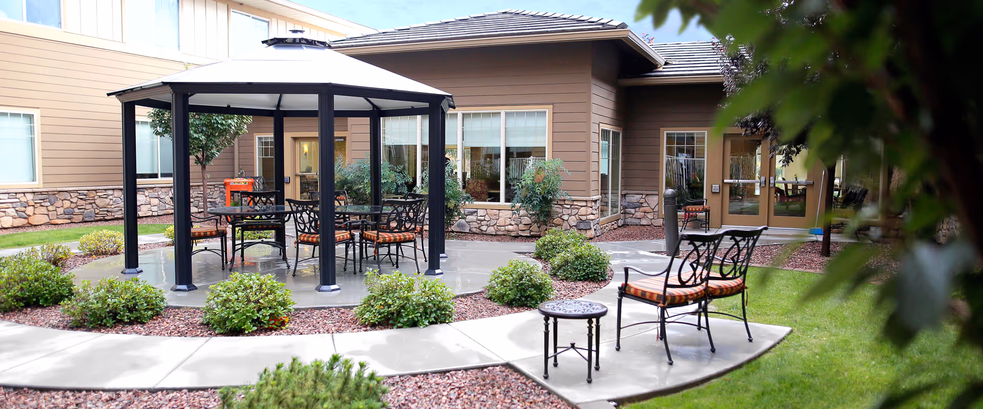 Outdoor patio area at a senior living facility featuring a gazebo with a table and chairs underneath, surrounded by small bushes and a curved concrete pathway. The building exterior has brown siding with stone accents and multiple windows.