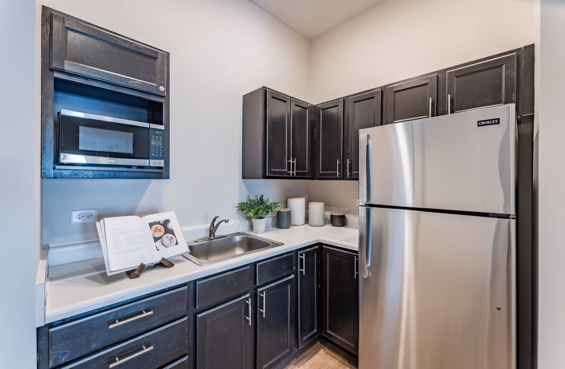 A modern kitchen corner featuring dark wood cabinets, a stainless steel refrigerator, a built-in microwave, a sink with a faucet, a cookbook on a stand, and decorative containers and a small plant on the countertop.