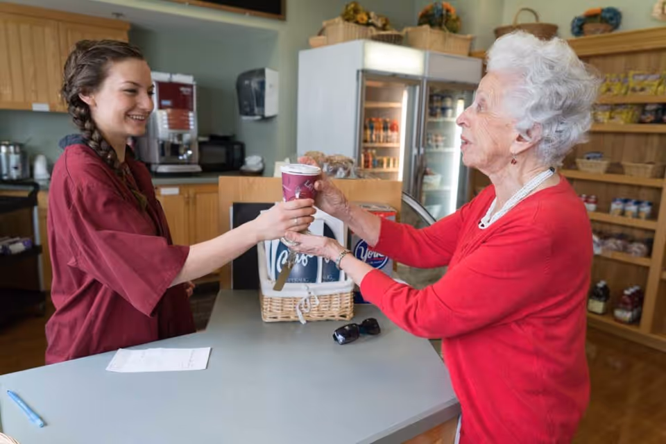 A young woman in a red uniform hands a cup of coffee to an elderly woman wearing a red sweater across a counter in a small shop or cafe area with shelves of snacks and a refrigerator in the background.
