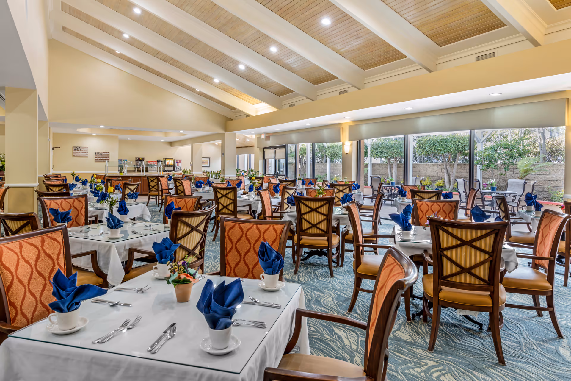 A spacious dining room with multiple tables set with white tablecloths, blue folded napkins in cups, silverware, and small flower arrangements. The room features large windows letting in natural light and a view of an outdoor patio area with greenery. The ceiling has wooden beams and recessed lighting.
