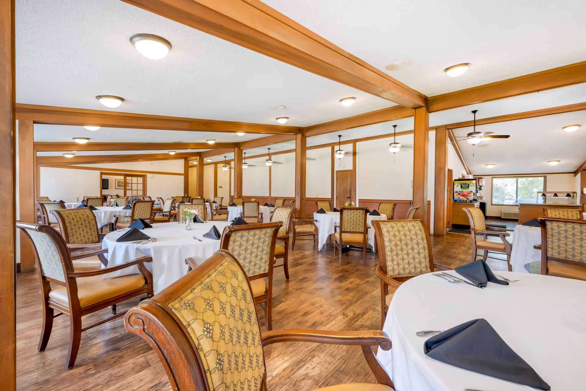 Spacious dining room with round tables covered in white tablecloths and wooden chairs arranged across a wood-floored room.