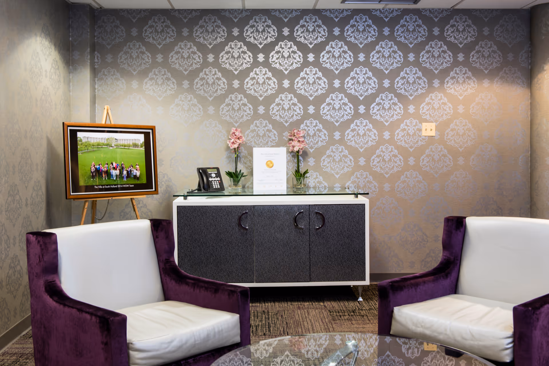 A small sitting area with two white and purple armchairs facing a glass coffee table. Behind the chairs is a cabinet with a telephone, two pink flower arrangements, and a framed photo on an easel. The wall has a decorative silver damask pattern wallpaper.