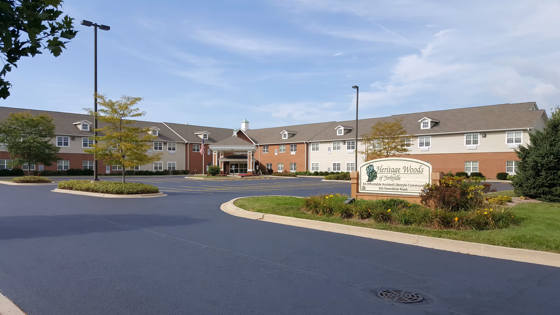 Exterior view of Heritage Woods of Yorkville, an assisted living community. The building is two stories with a combination of brick and light-colored siding. There is a covered entrance with columns, an American flag, and a parking lot with marked spaces. Trees and landscaped greenery surround the area under a partly cloudy sky.