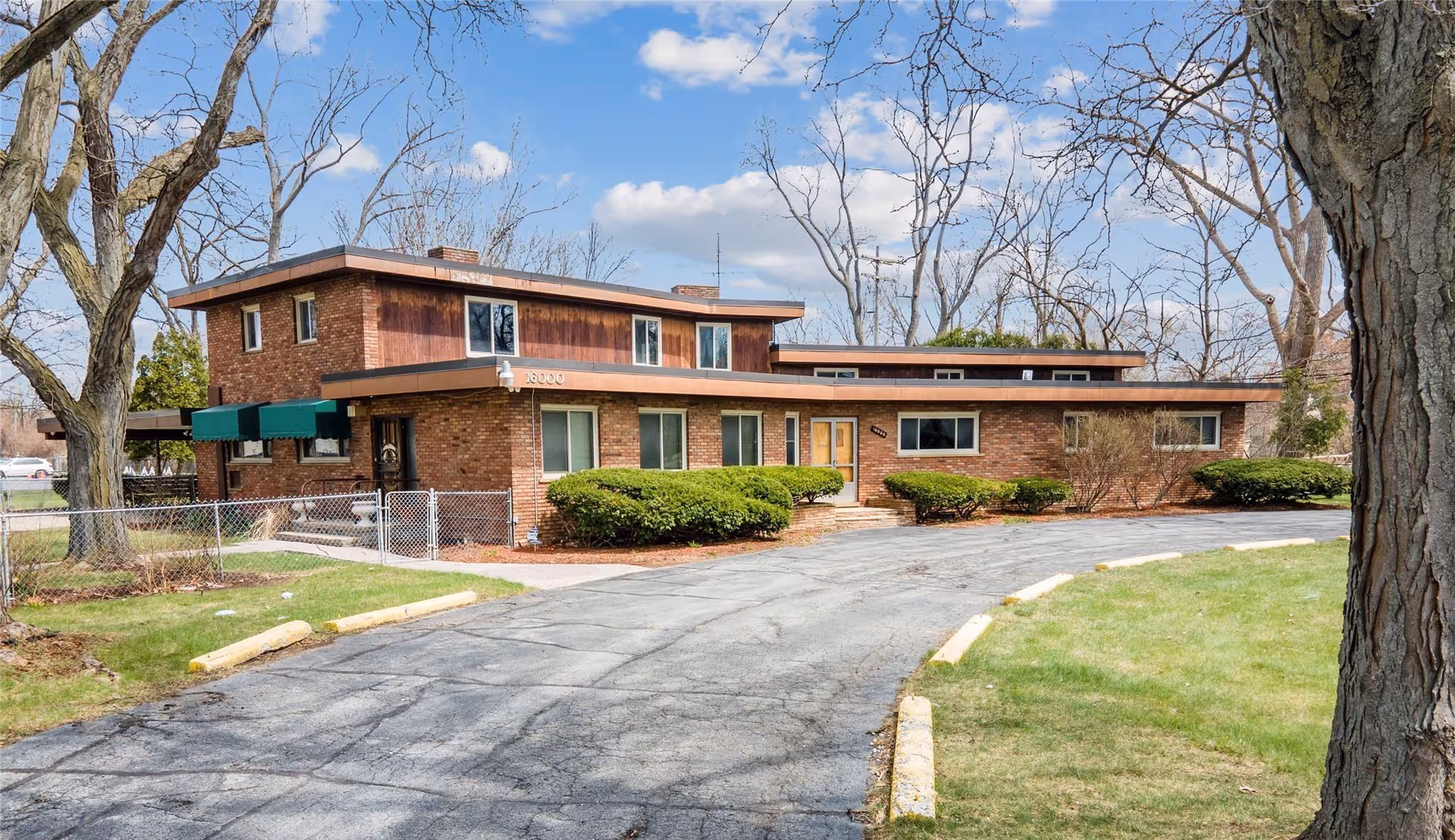 Exterior view of a two-story brick building with a flat roof, surrounded by leafless trees and green bushes. The building has several windows and a driveway leading up to the entrance. The sky is partly cloudy.