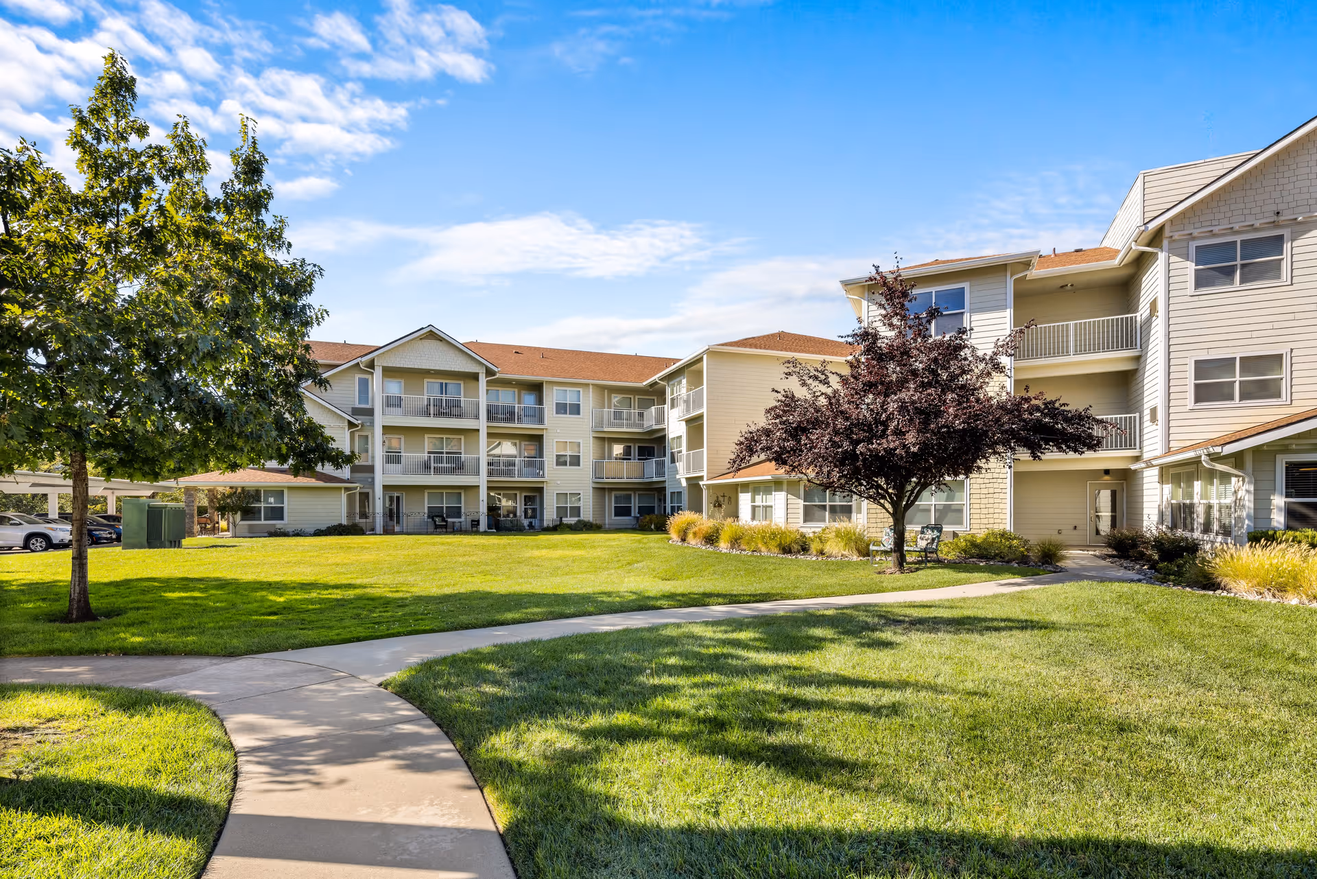 Outdoor view of Shasta Estates Senior Living showing a well-maintained grassy courtyard with a curved concrete pathway, trees, and a three-story residential building with balconies under a partly cloudy blue sky.