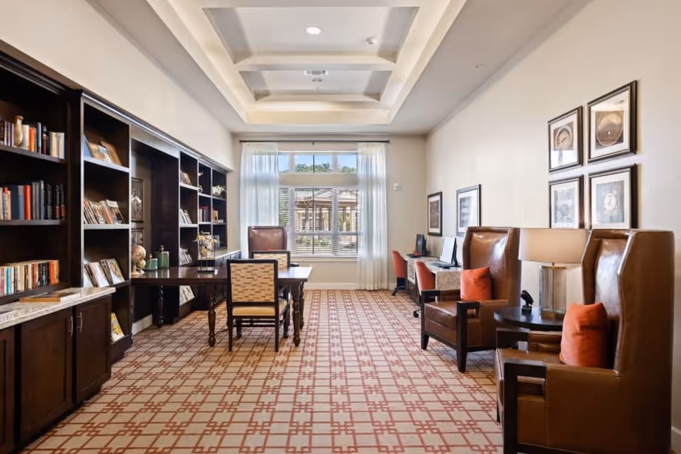 A bright communal library-style lounge with built-in bookshelves, tables and leather armchairs facing a large window.