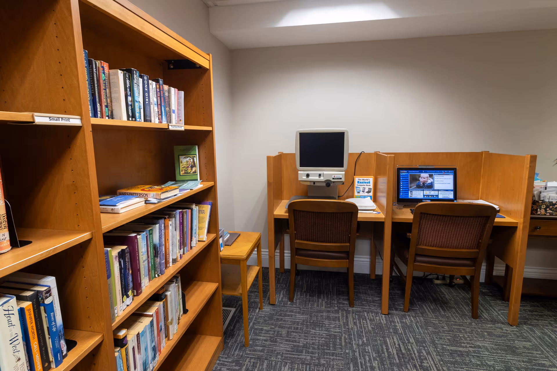 A small library or computer room with wooden bookshelves filled with books on the left side and two wooden computer desks with chairs on the right side. One desk has an older computer monitor, and the other has a laptop displaying a video call or news. The room has carpeted flooring and plain white walls.