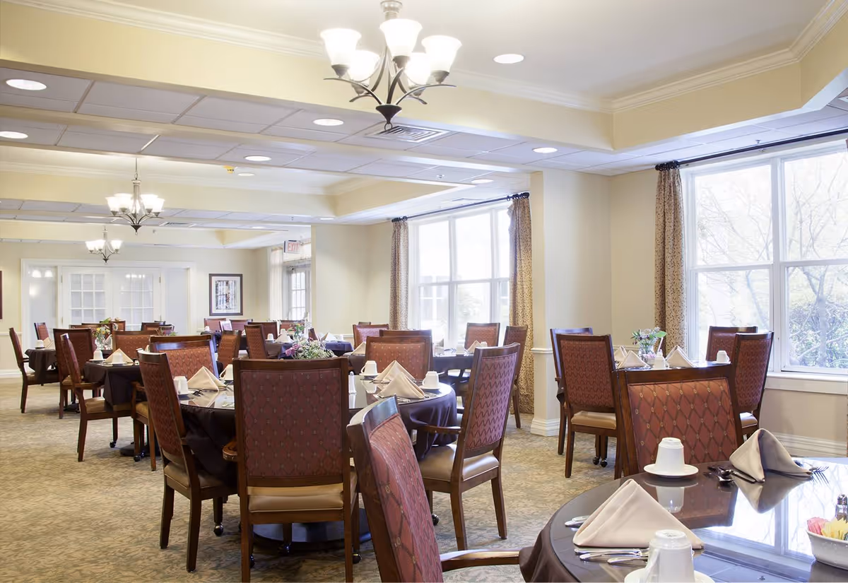 A bright and spacious dining room with multiple round tables covered in dark tablecloths, each set with folded napkins, cups, and silverware. The room has large windows with curtains allowing natural light to fill the space, beige walls, and chandeliers hanging from the ceiling. The chairs have patterned upholstery and wooden frames.