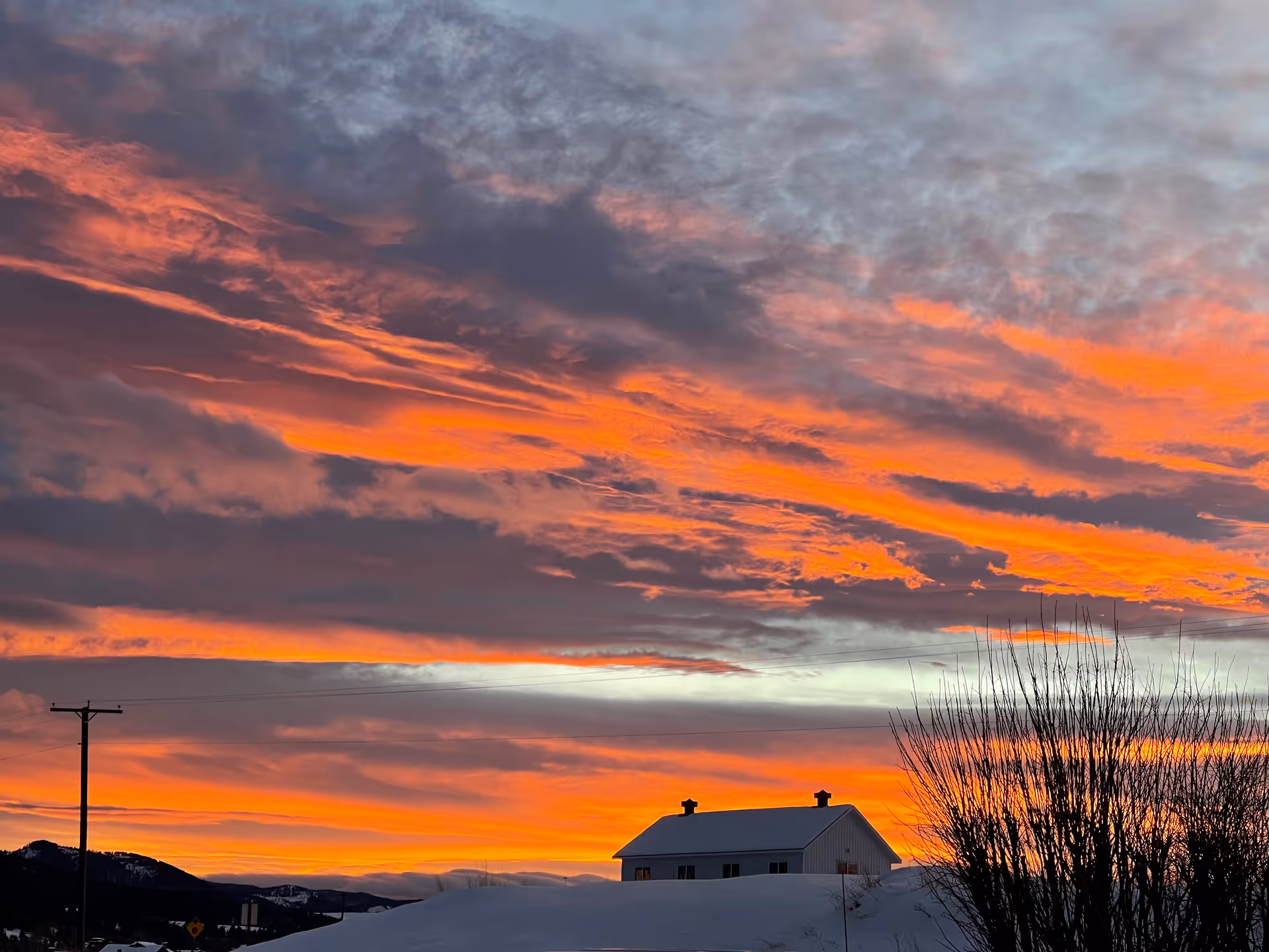 Snow-covered landscape at sunset with a silhouetted house, power pole, and bare trees beneath a fiery orange sky.