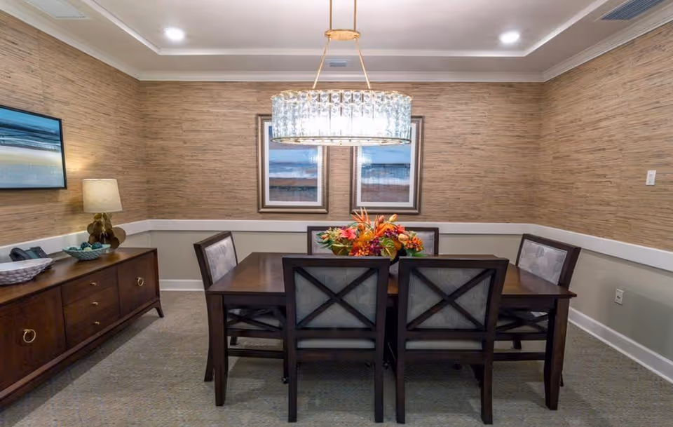 A dining room with a dark wooden table surrounded by six chairs with cushioned backs. A floral centerpiece decorates the table. The walls have a textured beige wallpaper with a white chair rail. Two framed landscape paintings hang on the far wall. A modern chandelier with a circular glass design hangs above the table. To the left, there is a wooden sideboard with a lamp, a telephone, and decorative bowls. The room has a carpeted floor and a tray ceiling with recessed lighting.