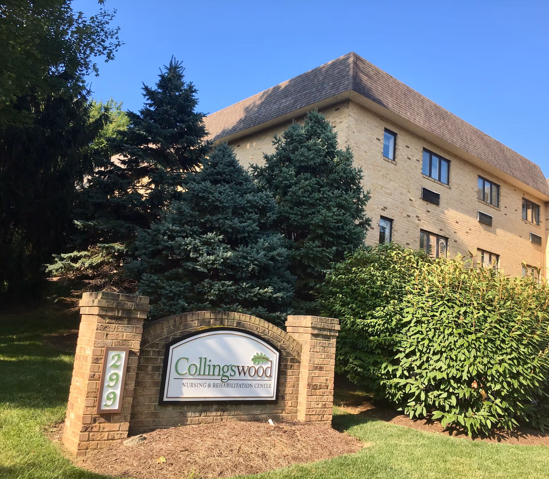 Exterior view of Collingswood Nursing & Rehabilitation Center building with a brick sign in front displaying the facility name and the number 299. The building is surrounded by green trees and bushes under a clear blue sky.