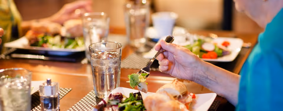 Close-up of people eating at a dining table with plates of salad, glasses of water, and a hand holding a fork.