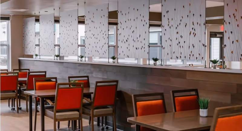 A dining area with several wooden tables and chairs featuring orange cushions. The room has large windows with decorative vertical blinds that have a leaf pattern. Small potted plants are placed on the tables and along the window ledge, creating a warm and inviting atmosphere.