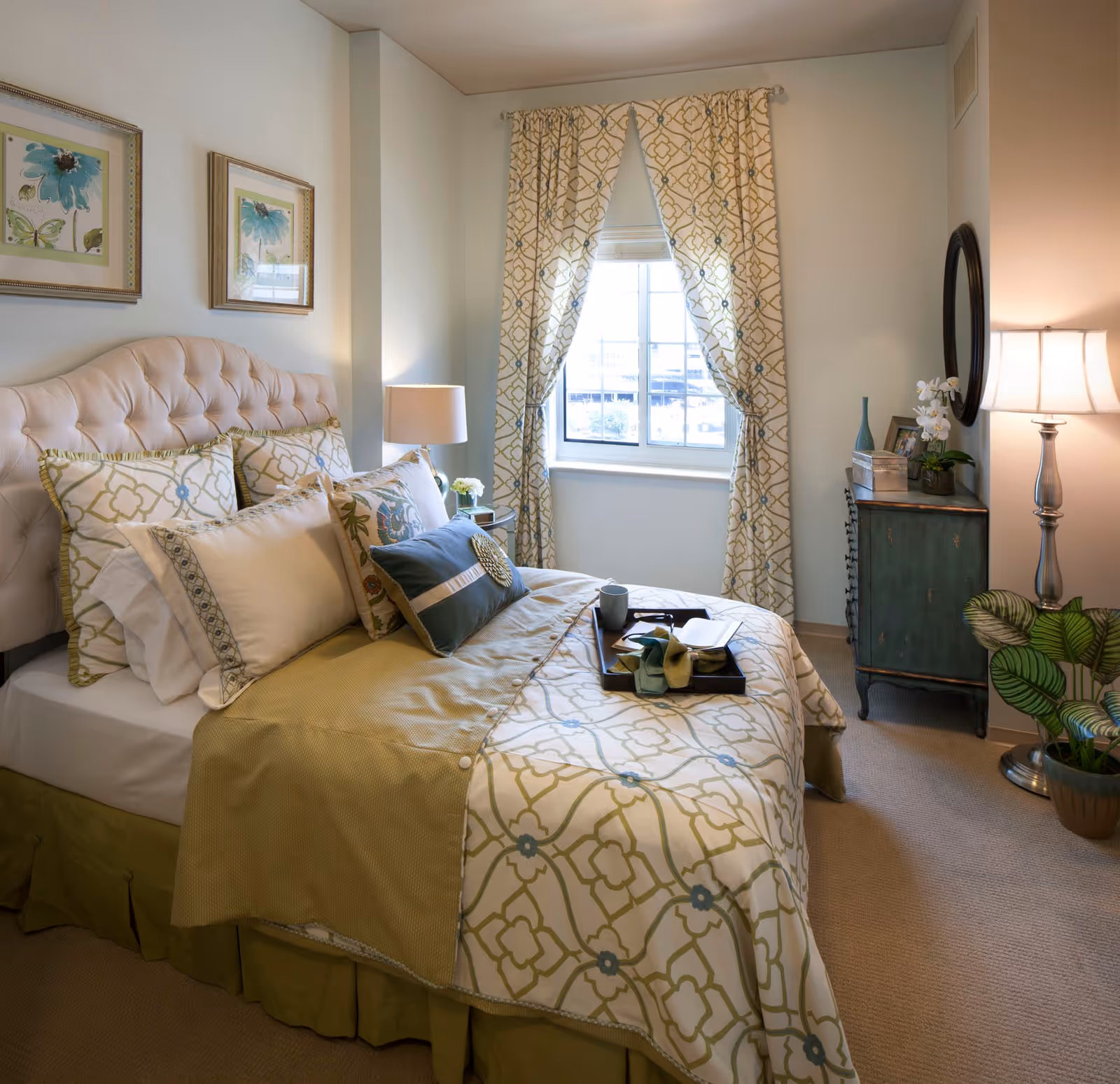 A neatly made bed with a tufted headboard in a bedroom. The bed has multiple pillows and a patterned bedspread in shades of green and cream. There is a window with patterned curtains behind the bed. A small green dresser with decorative items and a round mirror is on the right side of the room. A floor lamp and a potted plant are also visible in the corner.