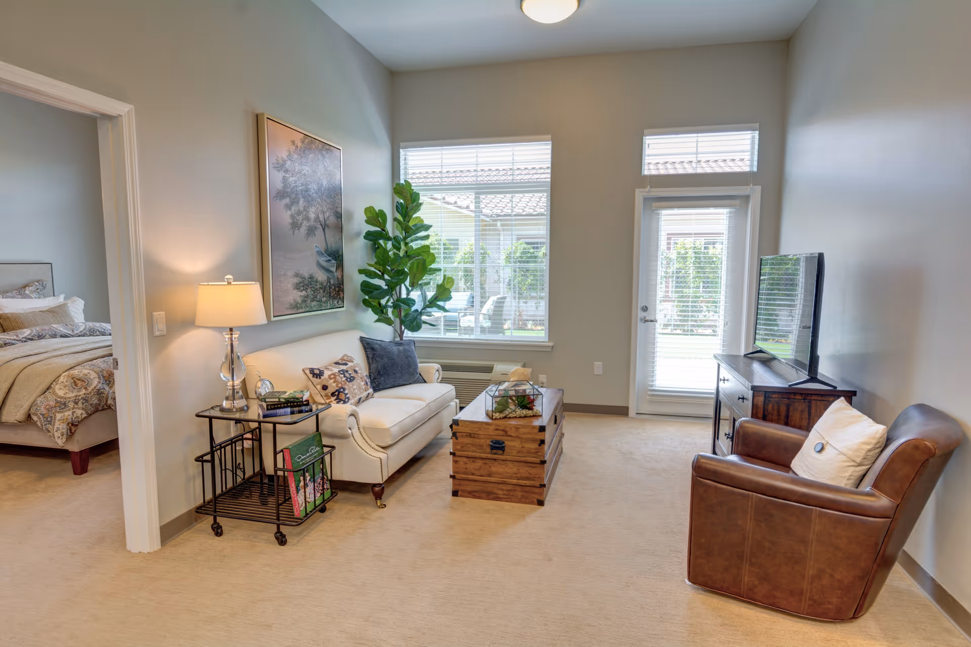 A cozy living room with a white sofa adorned with decorative pillows, a wooden chest used as a coffee table, a brown leather armchair with a white pillow, and a wooden TV stand with a flat-screen TV. A side table with a lamp and books is next to the sofa. Large windows and a glass door allow natural light to fill the room. A bedroom with a made bed is visible through an open doorway.