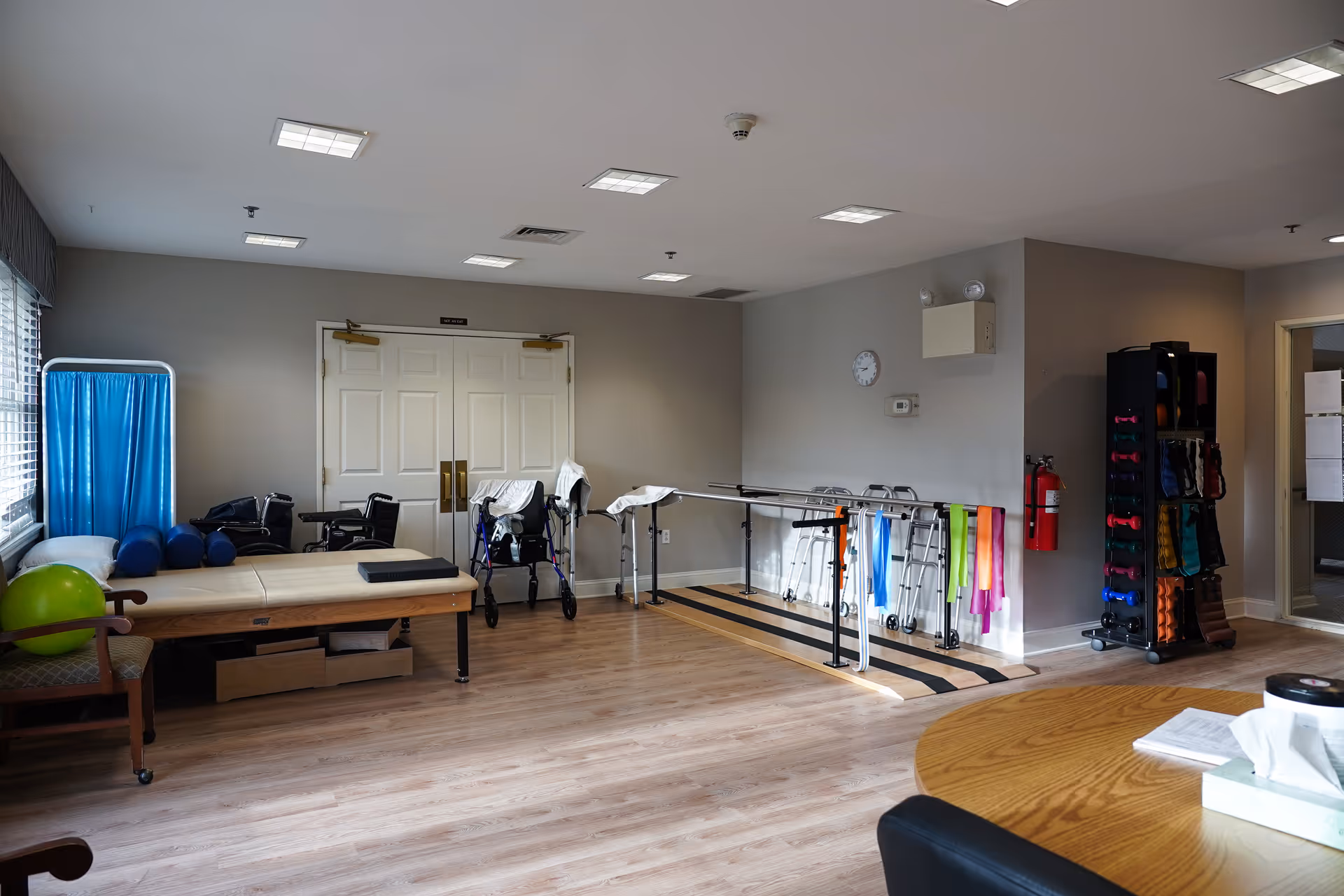 A rehabilitation or physical therapy room with parallel bars for walking exercises, several walkers and wheelchairs, a padded therapy table, exercise balls, resistance bands, and a rack of small dumbbells. The room has wood flooring, beige walls, and fluorescent ceiling lights.