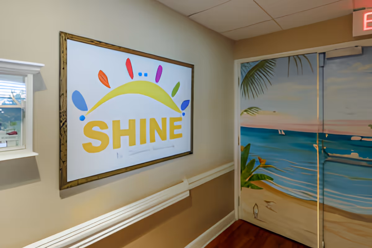 Interior hallway with beige walls and wooden floor. On the left wall, there is a framed sign with a colorful sun graphic and the word 'SHINE' in large yellow letters. On the right, double doors are painted with a beach scene featuring sand, ocean waves, palm leaves, and a sunset sky. An exit sign is visible above the doors.