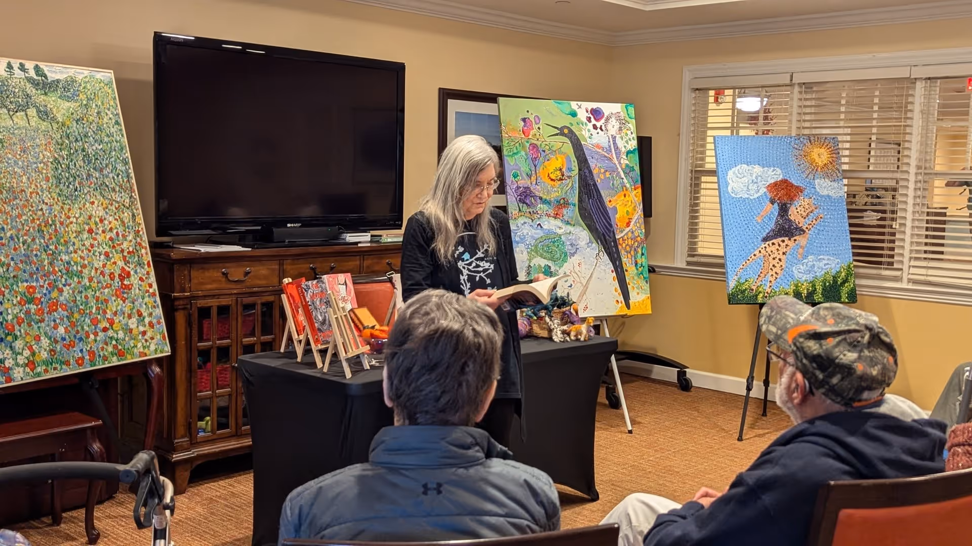 A woman with long gray hair is reading a book aloud to an audience seated in a room. Behind her are colorful paintings displayed on easels and a large flat-screen TV on a wooden cabinet. The room has beige walls and windows with blinds.