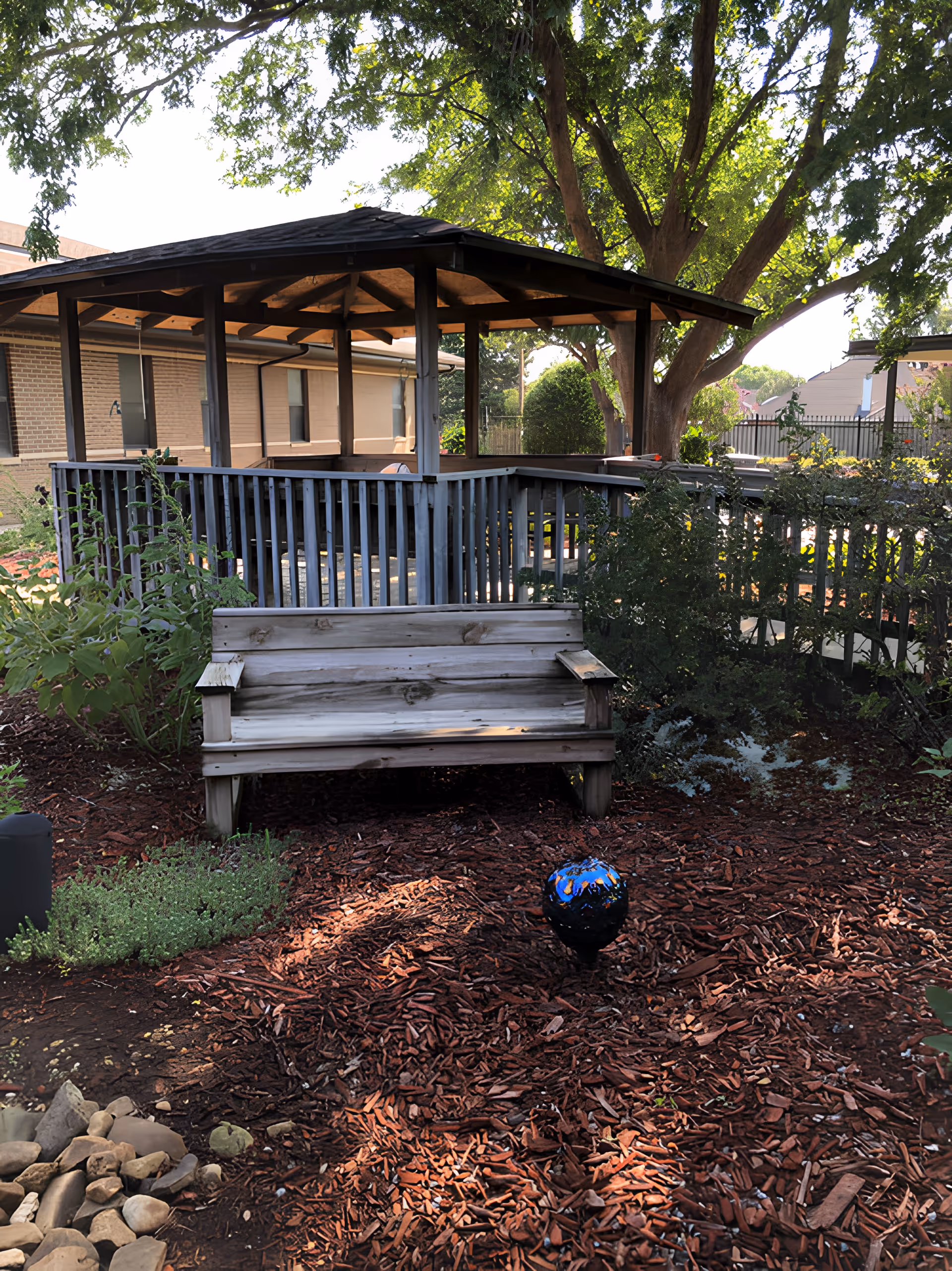 Outdoor garden area with a wooden bench in the foreground, a gazebo with a shingled roof and wooden railing behind it, surrounded by trees and plants. A building is visible in the background.