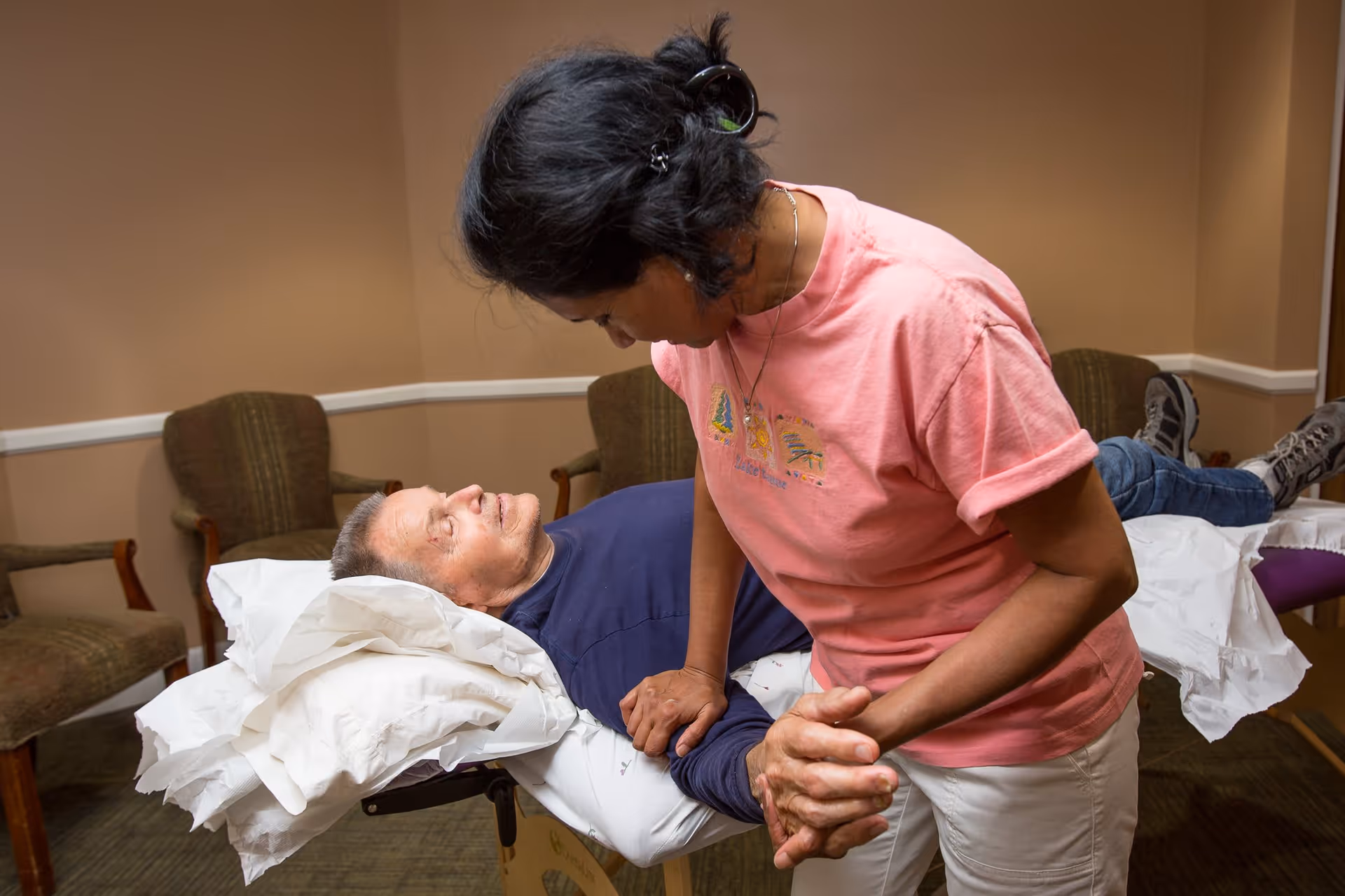 A woman in a pink shirt is assisting an elderly man lying on a padded table in a room with beige walls and several chairs. The woman is holding the man's hands and leaning over him, providing support or therapy.