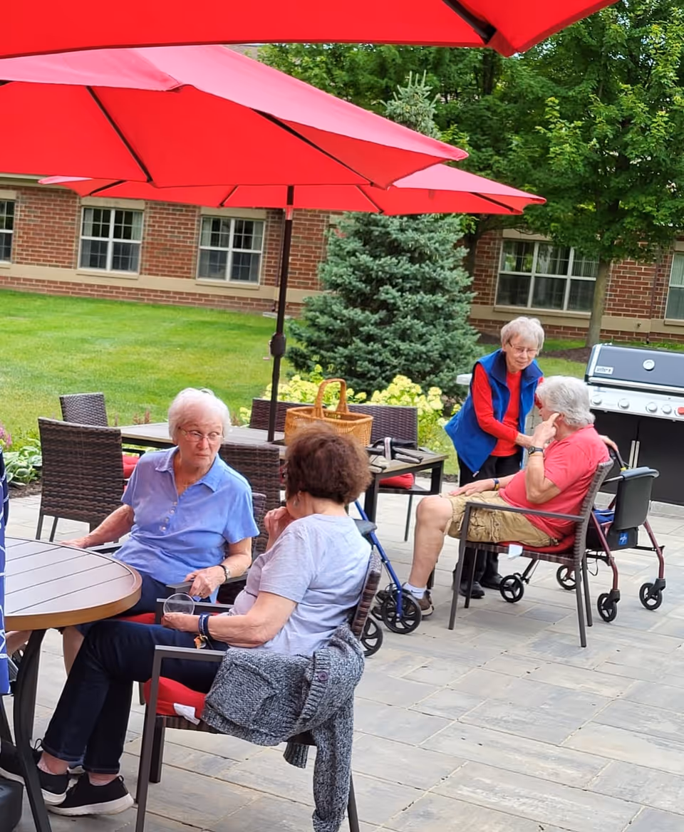 Four elderly people sitting and chatting outdoors on a patio with red umbrellas overhead. There is a green lawn, trees, and a brick building in the background. Two women are seated at a round table, while another woman is standing and talking to a man seated in a chair with a walker nearby. A barbecue grill is visible in the background.