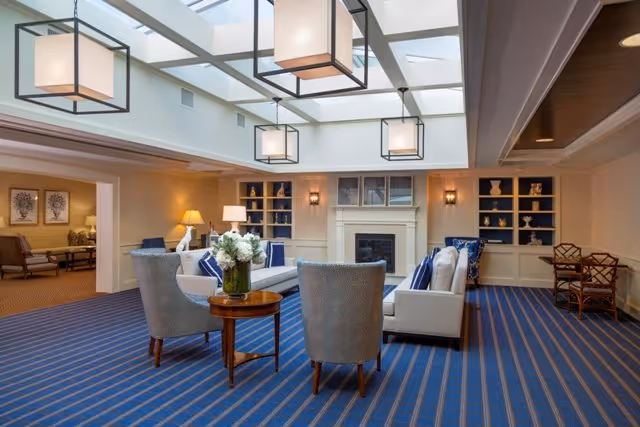 A spacious and well-lit living room area with a blue striped carpet, white sofas with blue and white cushions, two gray armchairs, a round wooden table with a flower arrangement, built-in shelves with decorative items, a fireplace, and large square pendant lights hanging from a glass ceiling.