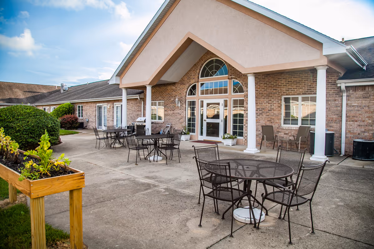 Outdoor patio area at Brookstone Estates of Mattoon South featuring metal tables and chairs on a concrete surface, with a raised wooden planter box containing green plants. The building has a brick exterior with white columns supporting a covered entrance and large windows and doors.