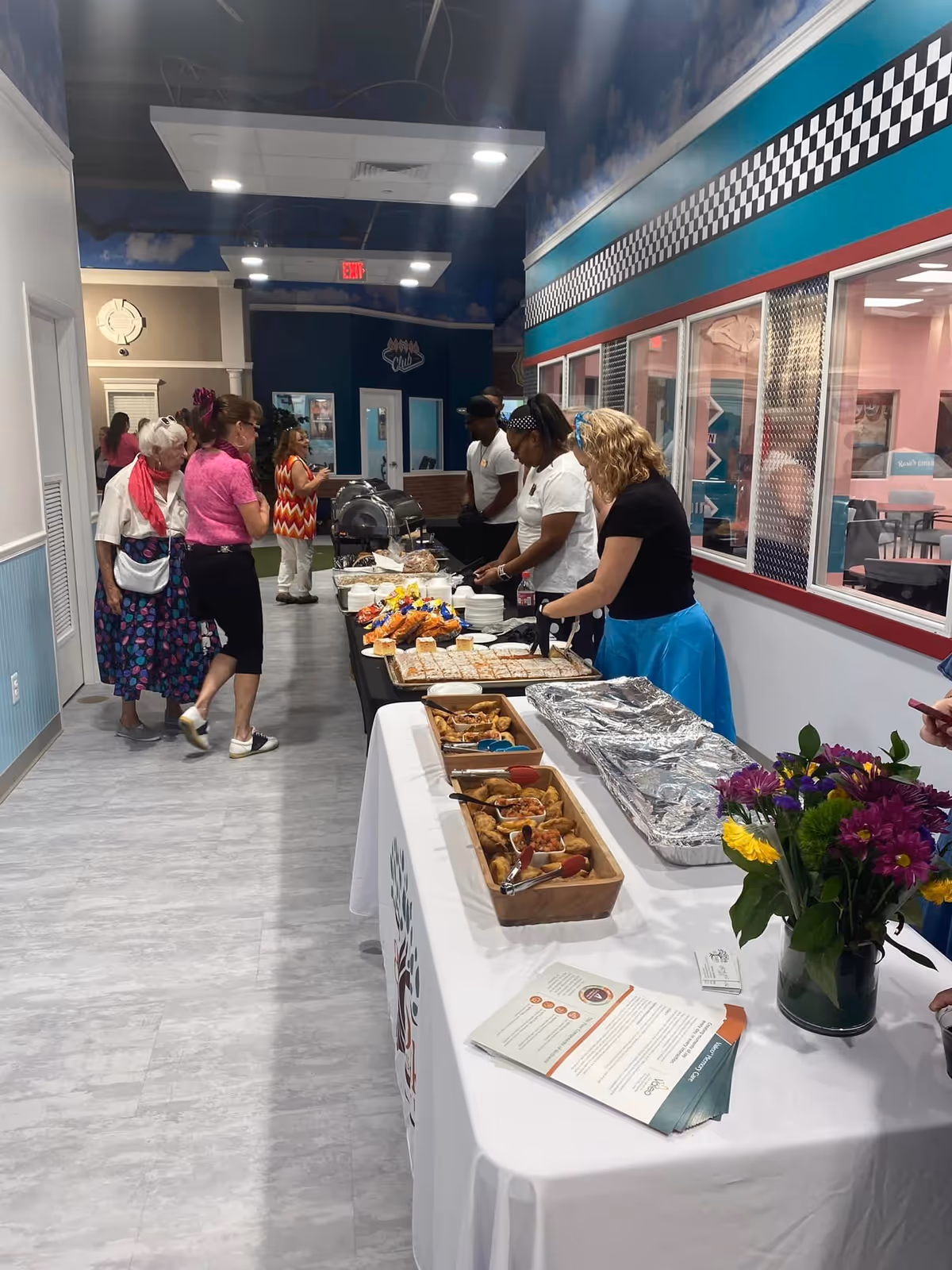 Indoor hallway with a buffet table set up along one side, featuring trays of food and flowers. Several people are standing near the buffet, serving themselves or waiting in line. The hallway has a checkered pattern decoration near the ceiling and windows looking into another room.