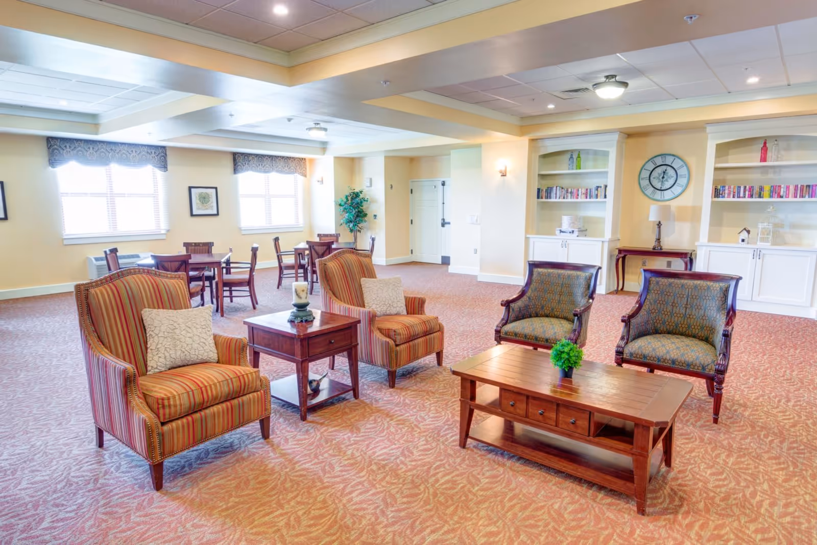 A spacious, well-lit common area with patterned carpet, featuring two striped armchairs with cushions, two patterned armchairs, a wooden coffee table with a small green plant, and a side table with a candle. In the background, there are several wooden tables and chairs near windows with valances, built-in white shelves with books and decorative items, a clock on the wall, and a small lamp on a side table.