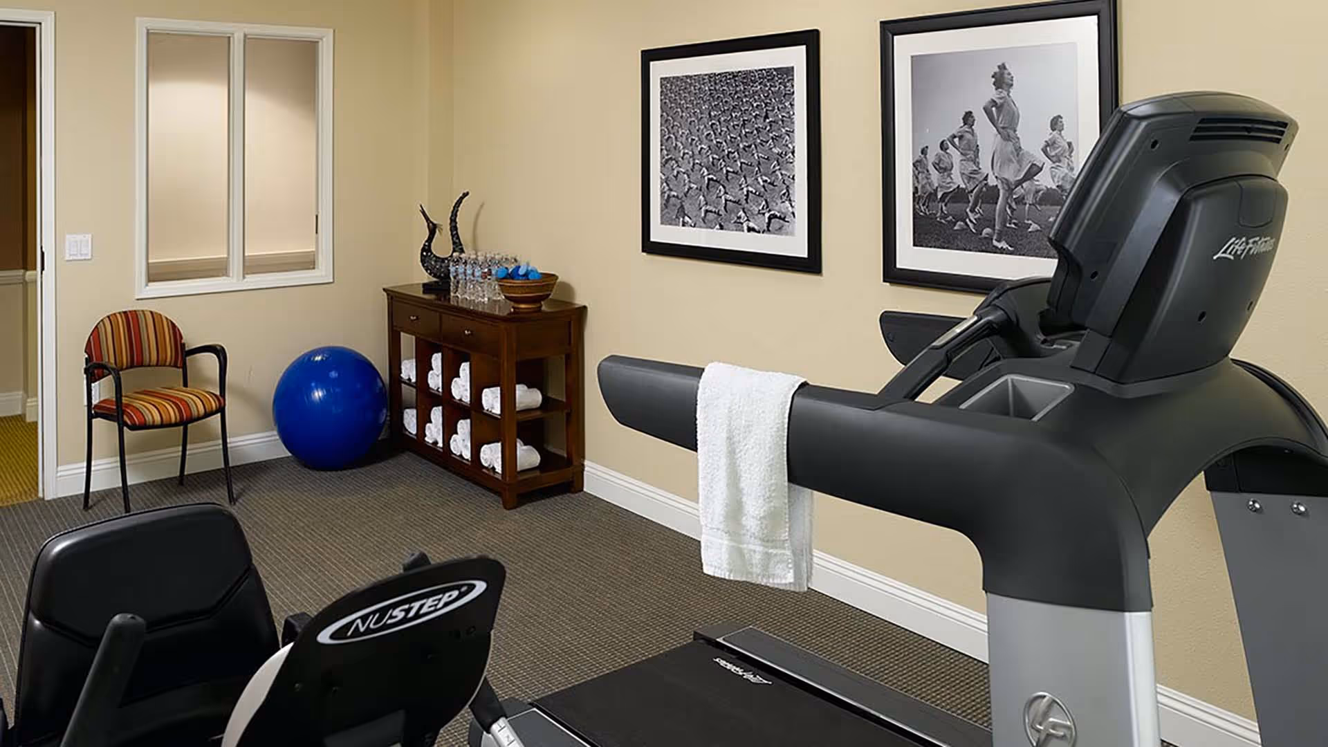 Interior view of a fitness room in a senior living facility with exercise equipment including a treadmill and a NuStep machine. A wooden shelf holds rolled white towels, water bottles, and small blue weights. Two black and white framed photos of people exercising hang on the beige wall. A striped chair and a large blue exercise ball are also visible.