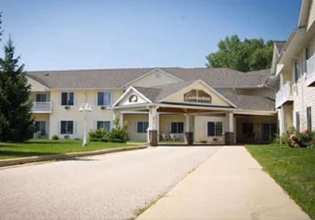 Exterior view of Charleston House Assisted Living facility showing a two-story building with a covered entrance, surrounded by green lawns and trees under a clear blue sky.