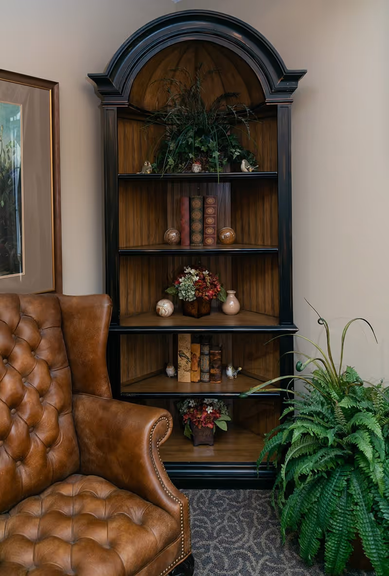 A cozy corner featuring a tufted brown leather armchair next to a tall, dark wooden corner shelf filled with decorative items such as books, small vases, and plants. A large green fern plant is placed on the floor to the right of the shelf, and a framed picture hangs on the wall to the left.