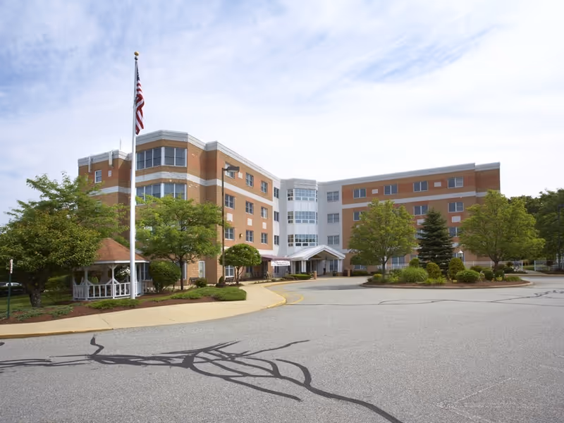 Exterior view of a multi-story healthcare facility building with a driveway in front, surrounded by trees and landscaping. An American flag is flying on a flagpole near a white gazebo on the left side of the image under a partly cloudy sky.