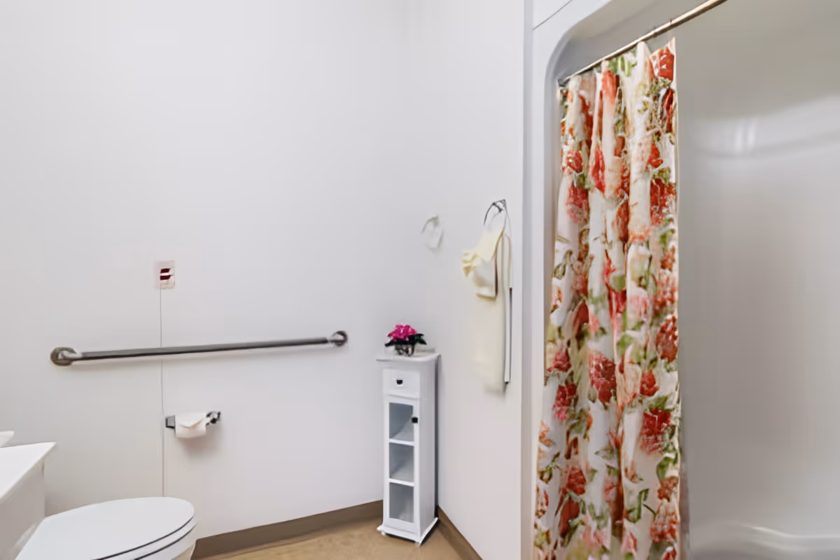 A clean bathroom with a white toilet, a metal grab bar on the wall, a toilet paper holder with a roll of toilet paper, a small white cabinet with a pink flower on top, a towel hanging on the wall, and a shower with a floral-patterned curtain.