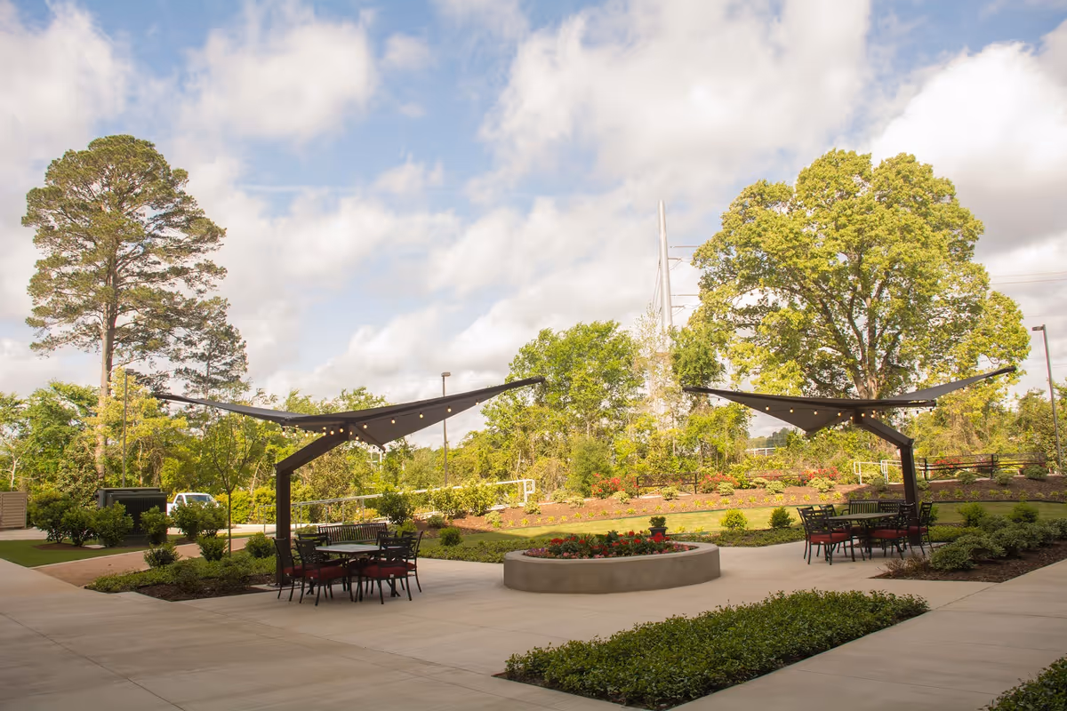 Outdoor patio area with two black metal tables and chairs under modern black shade structures with string lights. There is a circular concrete planter with flowers in the center, surrounded by landscaped greenery and trees under a partly cloudy sky.