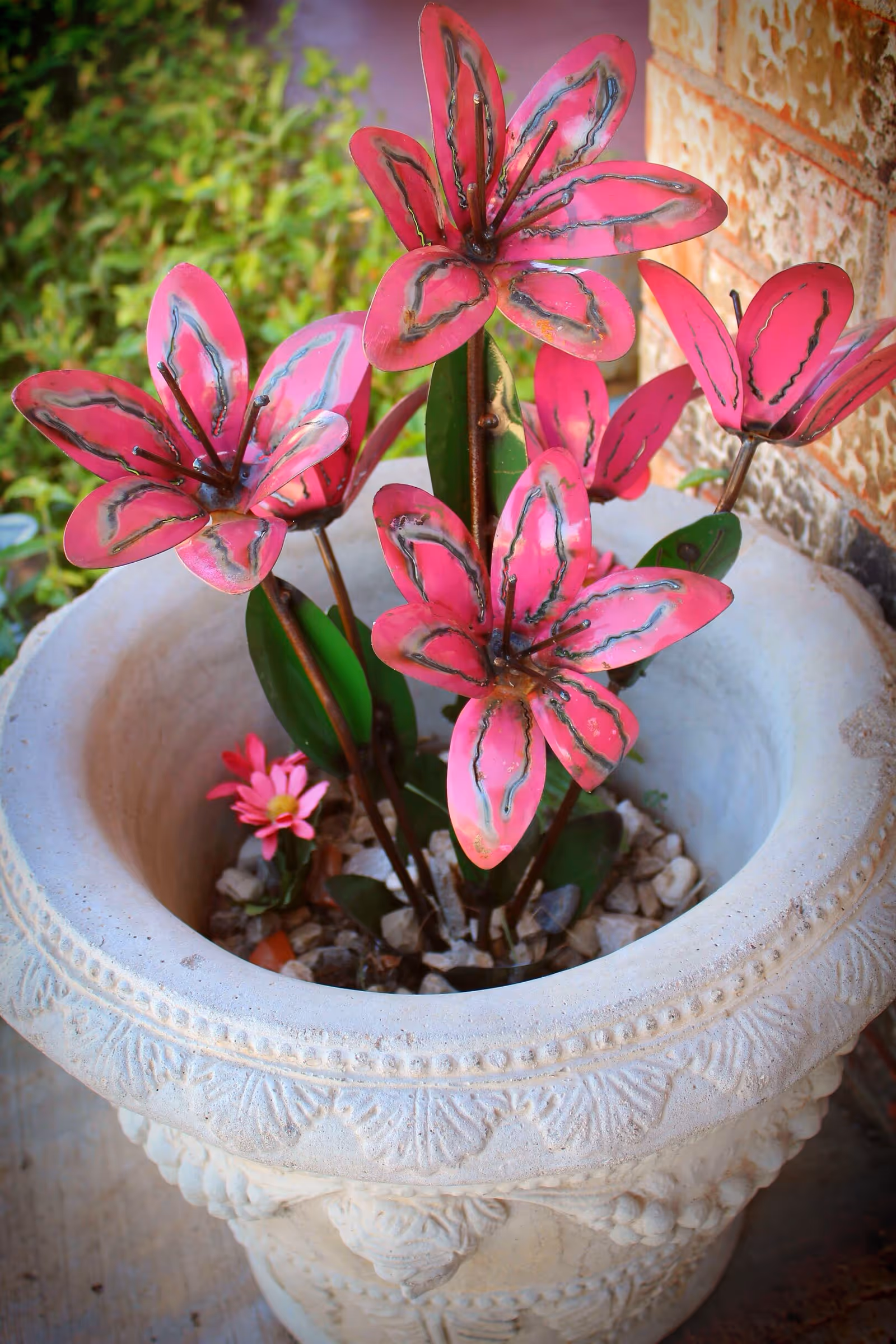 Stone planter holding pink metal flower sculptures and pebbles placed outdoors near a brick wall.