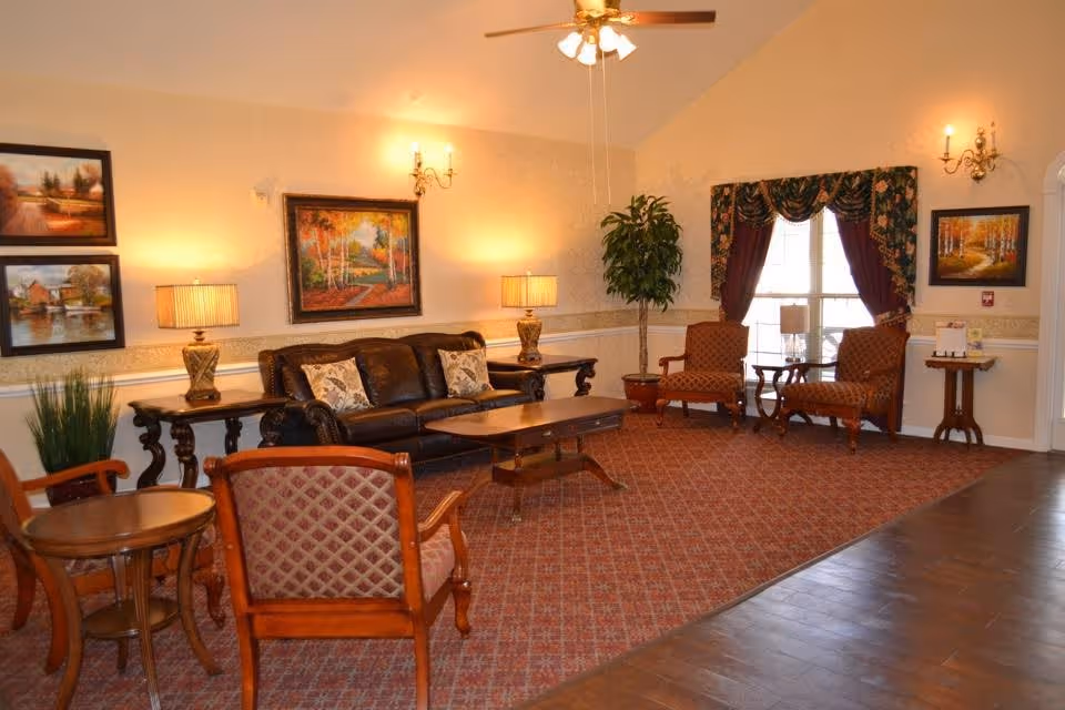 A cozy living room area with a brown leather sofa, two patterned armchairs, wooden coffee and side tables, two table lamps, a potted plant, and framed landscape paintings on the walls. The room has a patterned carpet, a ceiling fan with lights, and a window with floral curtains.