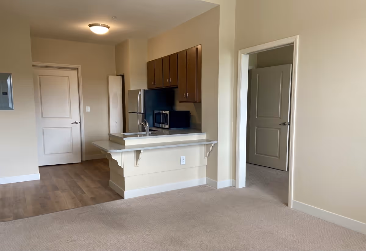Interior view of a senior living facility apartment showing a small kitchen area with wooden cabinets, a refrigerator, microwave, and a sink with a countertop extending into the living space. The floor transitions from wood in the kitchen to carpet in the living area. There are two open doors leading to other rooms.