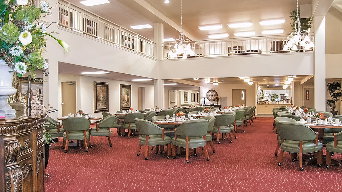 Spacious senior living dining room with multiple round tables set with white cups and plates, surrounded by green cushioned chairs on a red carpet. The room has a high ceiling with a balcony railing above, chandeliers, and decorative plants and paintings on the walls.