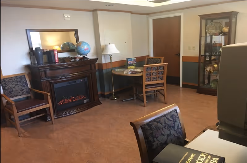 A cozy room with wooden furniture including chairs with floral upholstery, a round table, a floor lamp, and a wooden electric fireplace with a mirror and globe on top. There is a glass display cabinet with decorative items and a closed wooden door in the background.