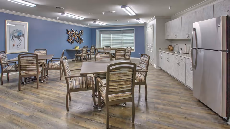 A dining area with several tables and cushioned chairs arranged on a wood floor. The walls are painted blue and decorated with a framed picture of a bird and a metal wall sculpture. On the right side, there is a kitchenette with white cabinets, a countertop, a sink, and a stainless steel refrigerator. A large window with blinds is visible at the back of the room.