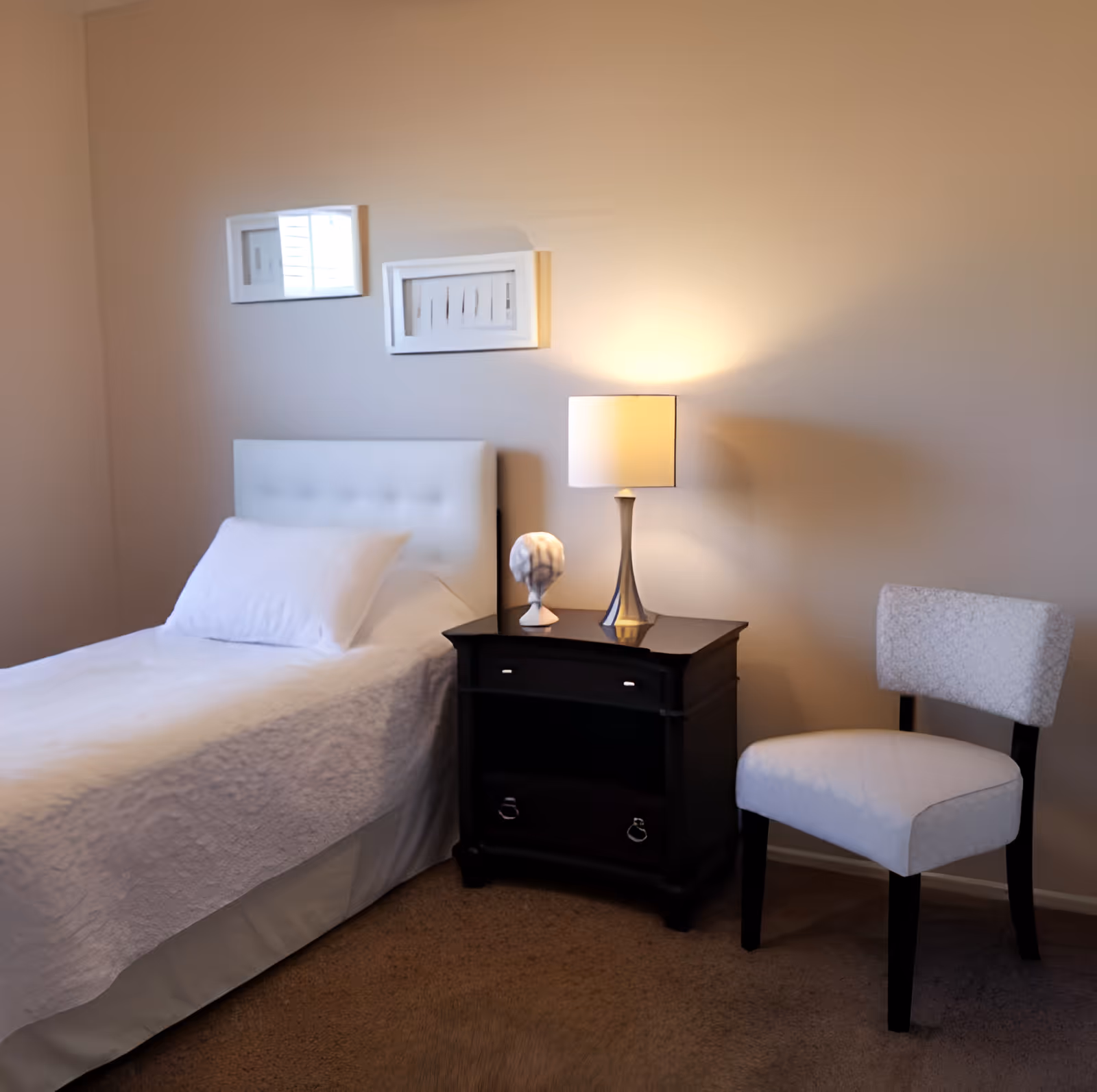 A simple bedroom with a single bed, a dark nightstand holding a lamp and decorative object, and a white upholstered chair against a beige wall.