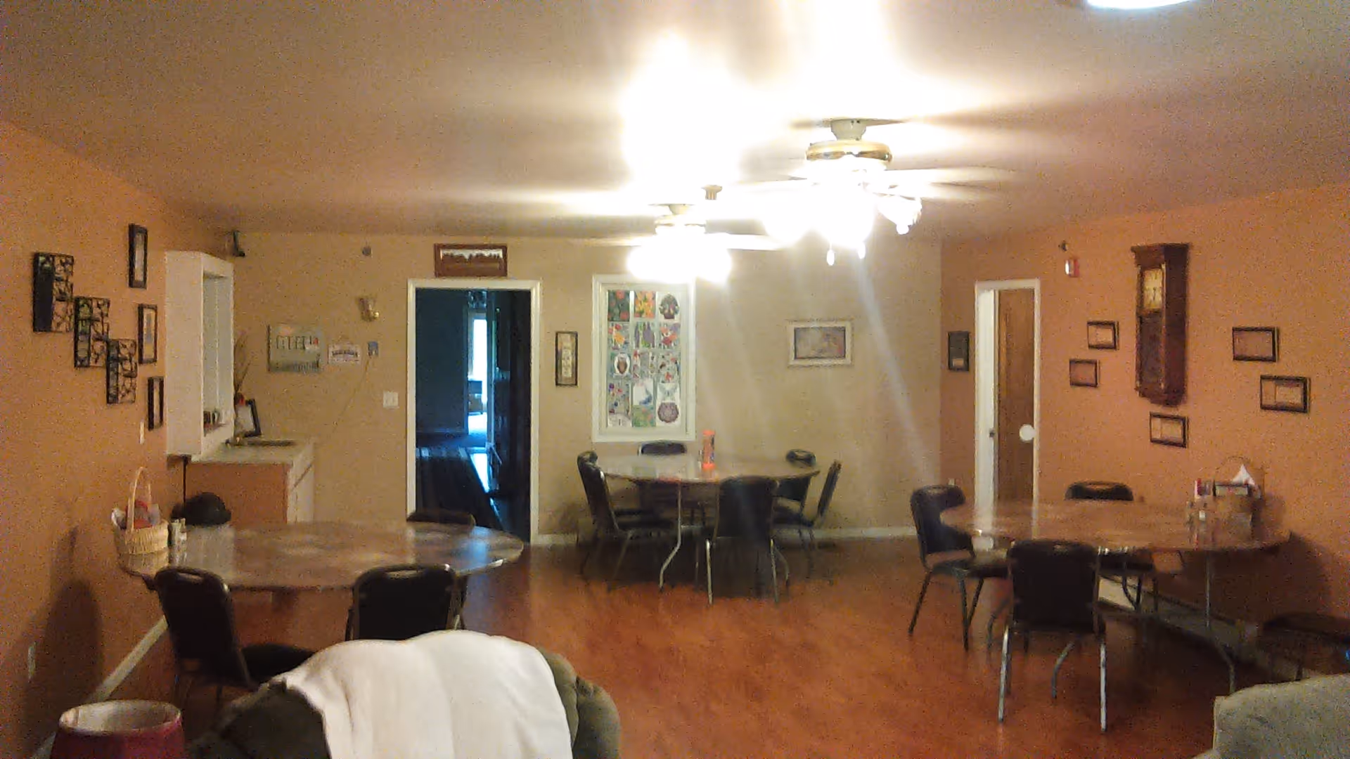 Interior view of a common area with multiple round tables and chairs arranged around the room. The walls are decorated with framed pictures and a wall clock. The floor is wooden, and ceiling fans with lights are mounted on the ceiling. There are doorways leading to other rooms.
