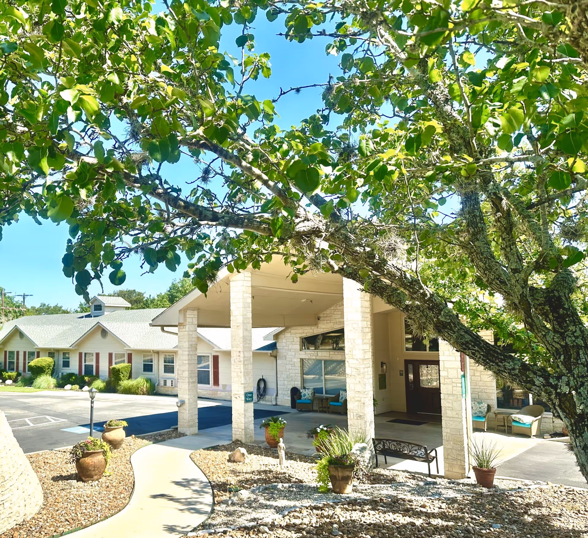 Entrance of Heritage Place of Boerne senior living facility with a covered driveway, stone pillars, outdoor seating, potted plants, and a large tree with green leaves in the foreground under a clear blue sky.