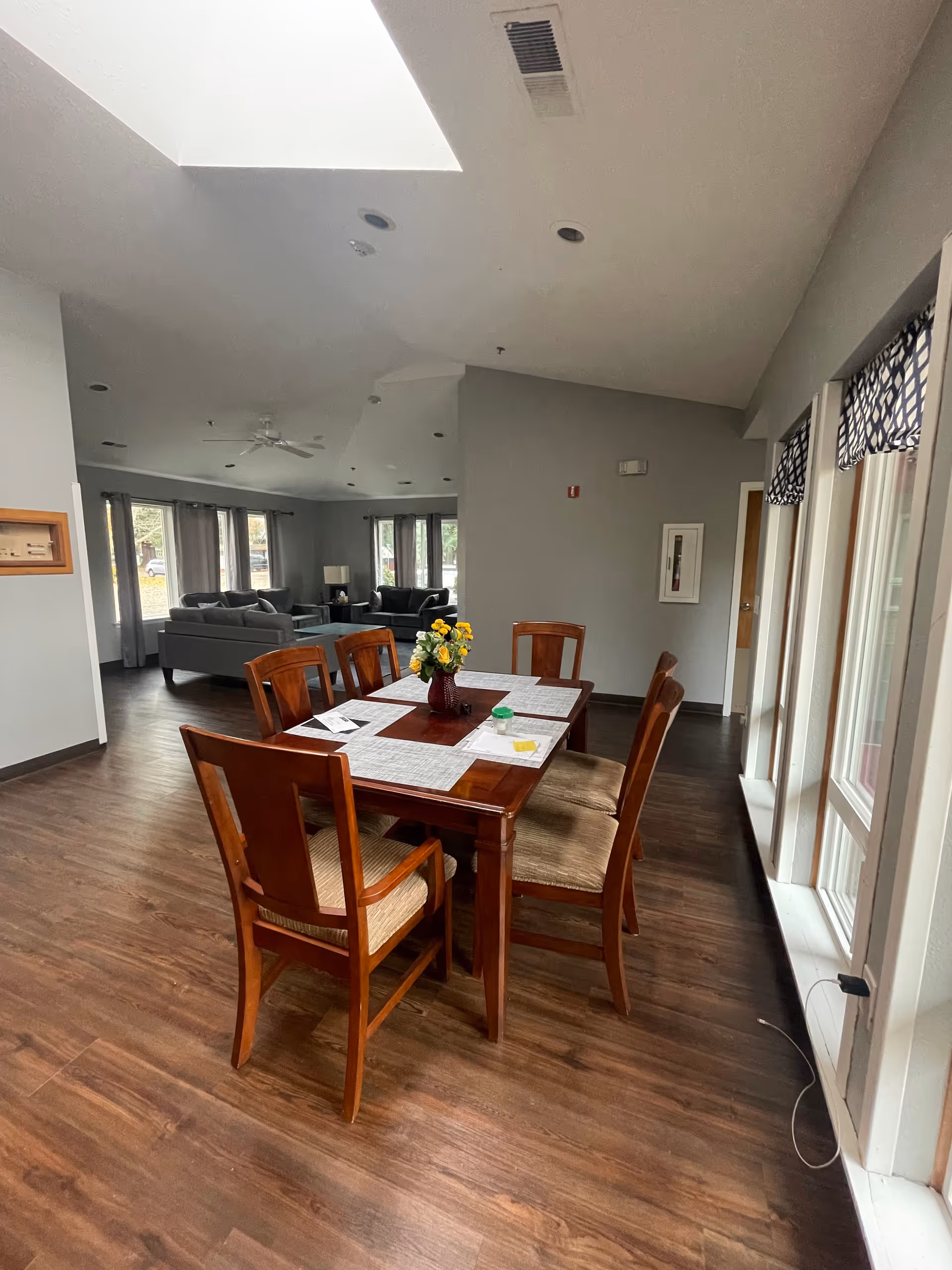 Interior dining area with a wooden table and six chairs on a wood floor, a vase of flowers on the table, and a living room seating area visible in the background.